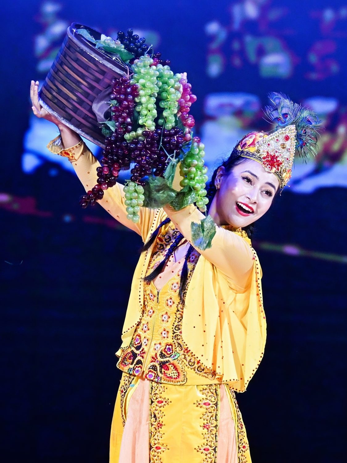 古扎丽阿衣舞台演出《葡萄熟了》,肩扛葡萄果篮,黄色艾德莱斯纹样演出服与孔雀翎冠